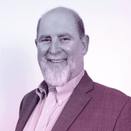 A middle-aged man with a gray beard smiles at the camera, wearing a suit jacket and collared shirt, against a plain light background.